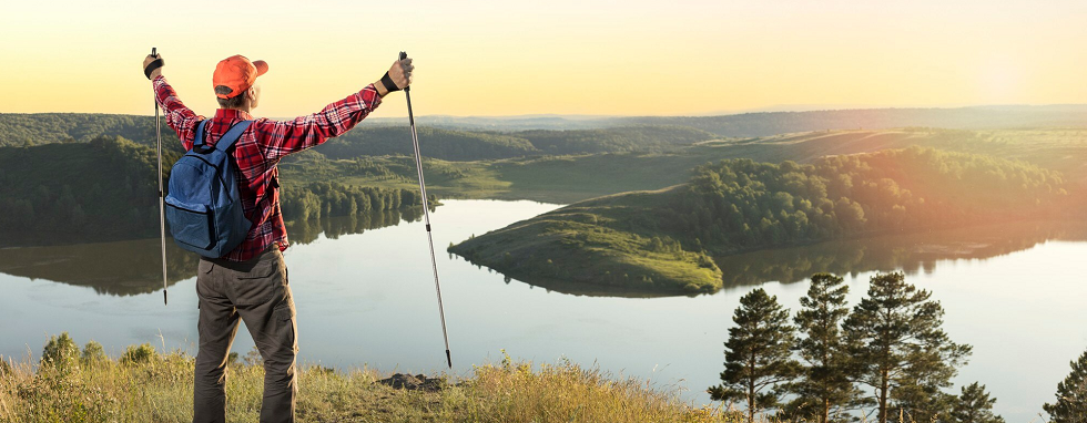 wyprawa trekkingowa - mężczyzna z plecakiem i kijkami podziwia widok na zbiornik wodny i zachód słońca